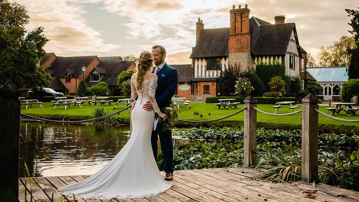 Bride and groom embrace on a wooden deck by the water at The Moat House, with lily pads, picnic benches, and the historic building in the background.