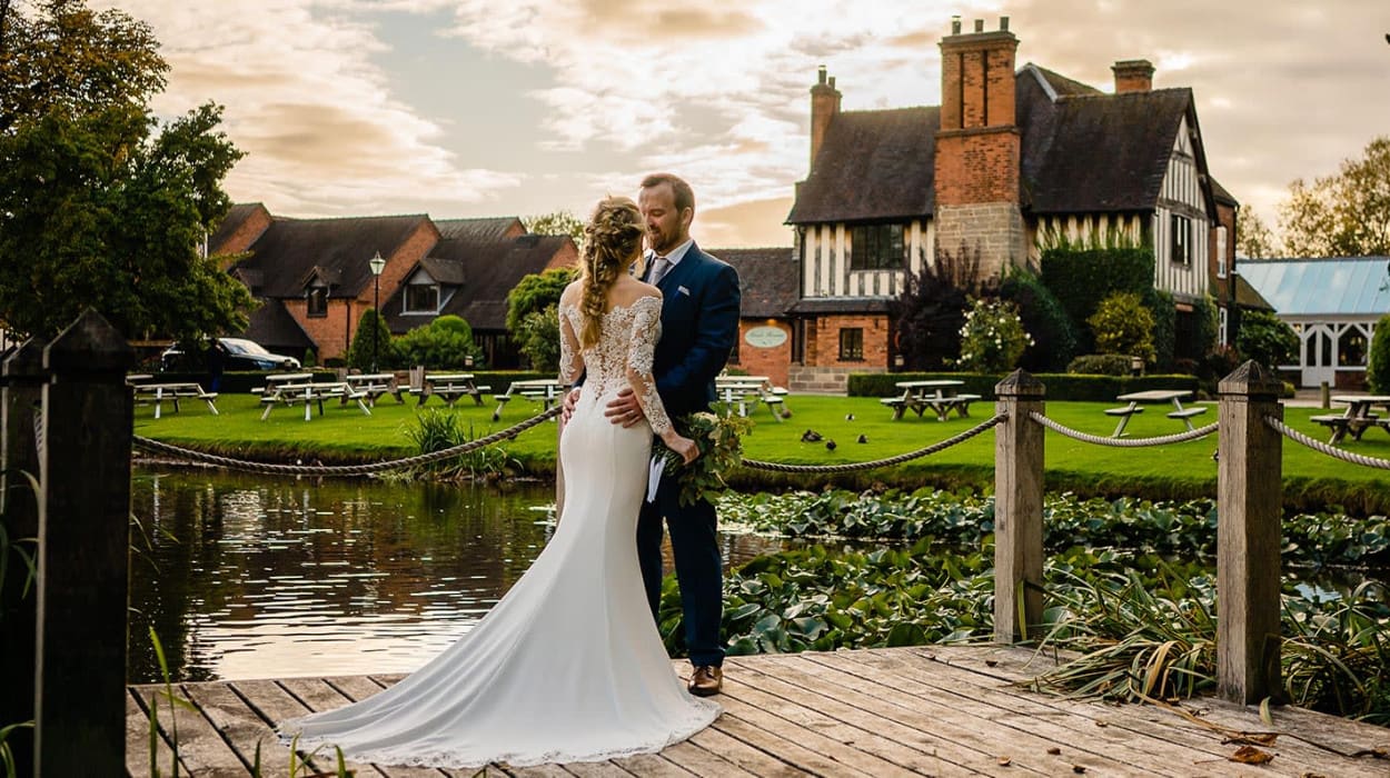 Bride and groom embrace on a wooden deck by the water at The Moat House, with lily pads, picnic benches, and the historic building in the background.