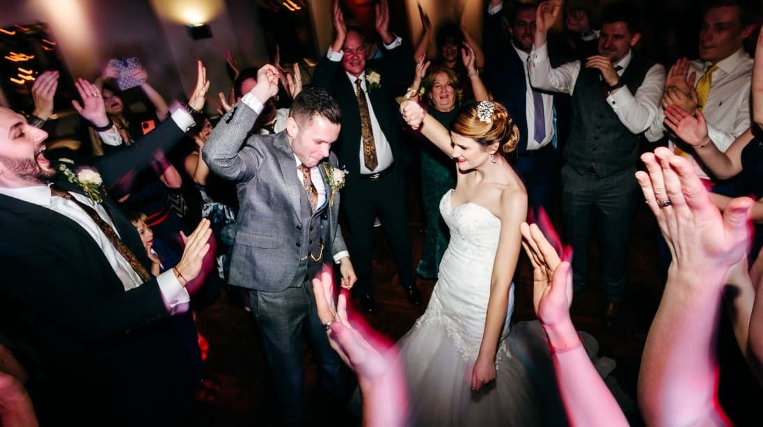 Alt-Text Generator Assistant said:  Bride and groom celebrate on the dance floor at The Moat House, surrounded by cheering guests with hands raised during a lively wedding reception.