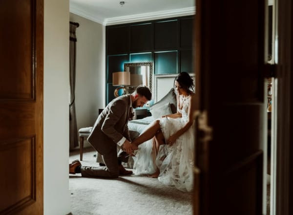 Groom kneels to help bride with her shoe in an elegant room at The Moat House, capturing an intimate wedding day moment before the ceremony.