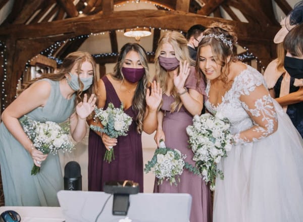 Bride and bridesmaids wearing face masks wave at a screen during a virtual wedding moment at The Moat House, holding bouquets in a rustic ceremony room.