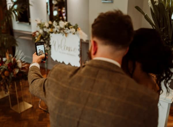 Couple takes a selfie together at a wedding, standing in front of a floral welcome sign and elegant decor inside a warmly lit venue.