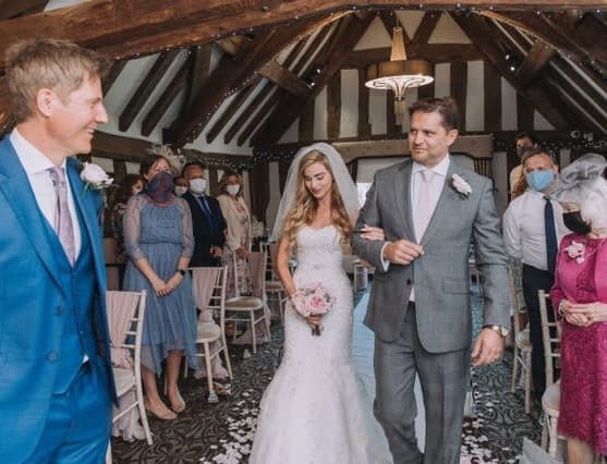Bride walks down the aisle with her father at The Moat House, as guests look on in a rustic ceremony room with exposed beams and petal-strewn carpet.