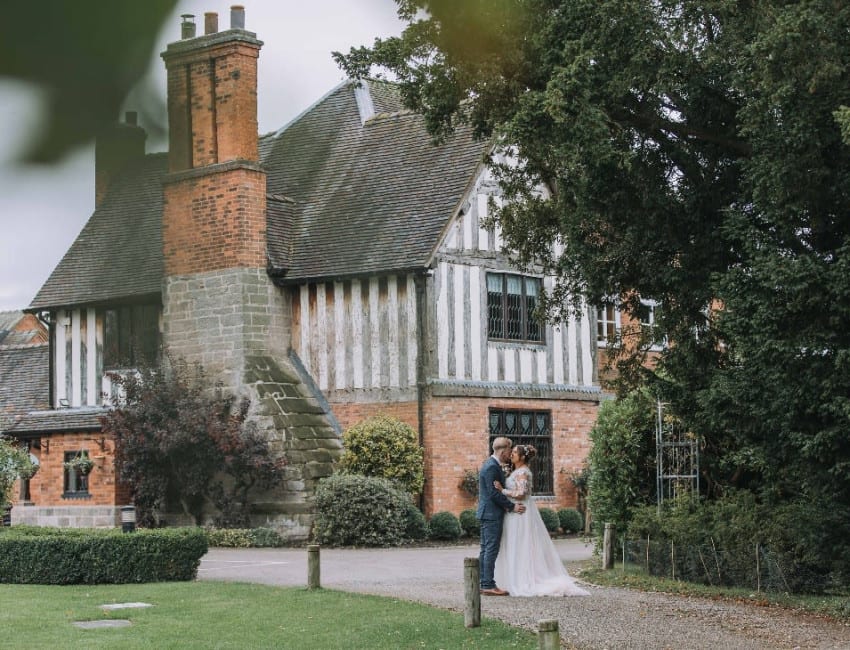 Bride and groom embrace outside a historic timber-framed building surrounded by lush greenery and gardens on their wedding day.
