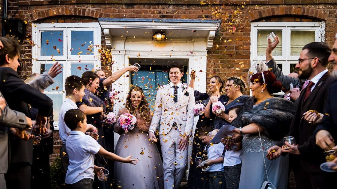 Bride and groom walk through a joyful crowd of guests throwing confetti outside a brick building during their wedding celebration.