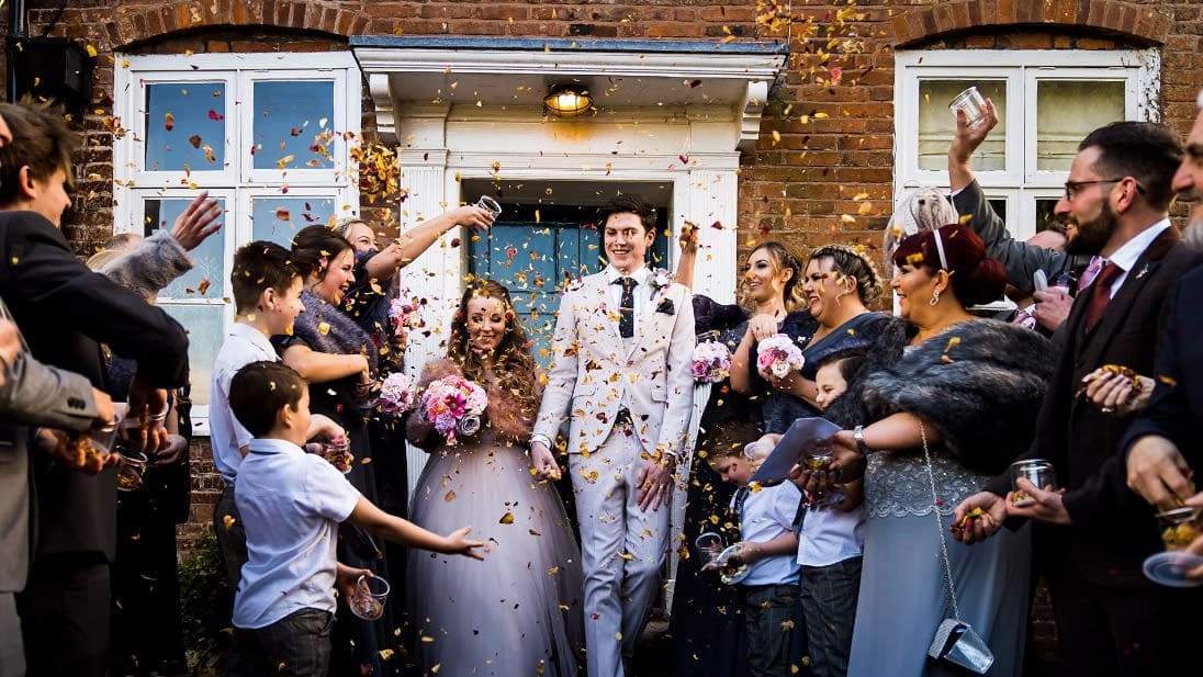 Bride and groom walk through a joyful crowd of guests throwing confetti outside a brick building during their wedding celebration.