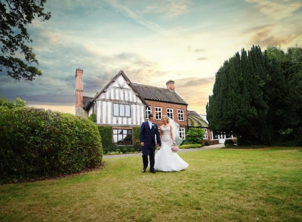 Bride and groom walk hand in hand on the lawn in front of The Moat House at sunset, with the historic timber-framed building and manicured gardens behind them.