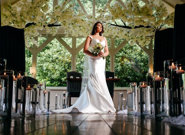 Bride stands at the altar in a white gown, holding a bouquet, surrounded by candlelit vases and white floral arches at The Moat House ceremony room.