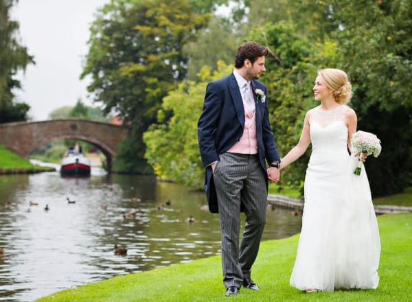 Bride and groom walk hand in hand along the canal at The Moat House, with ducks in the water and a boat approaching a brick bridge in the background.
