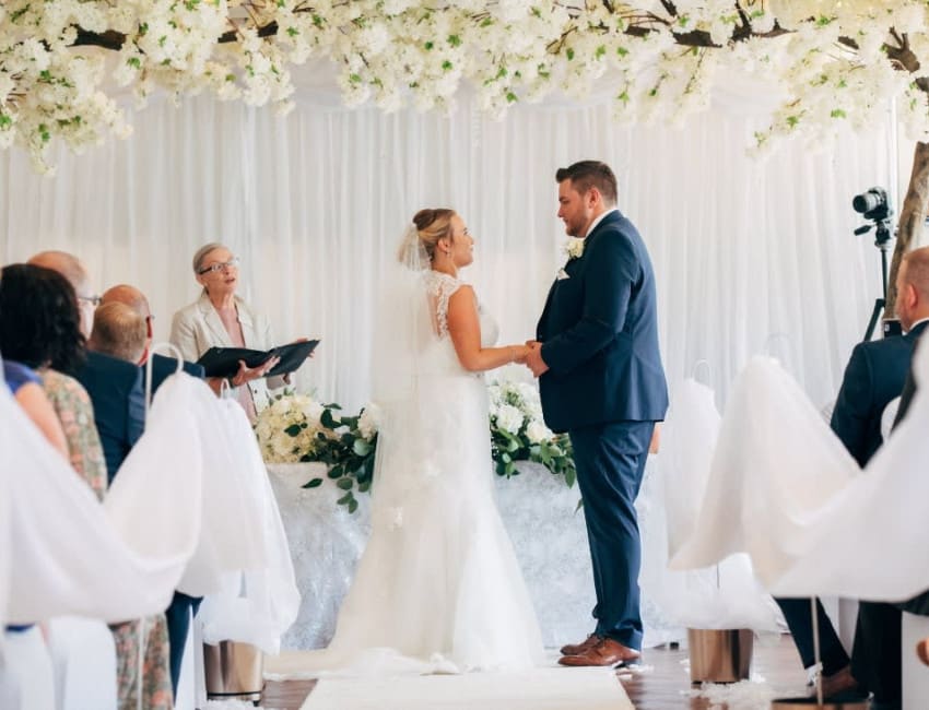 Photo-07-11-2019-08-32-15-1Bride and groom exchanging vows under white floral decor during a wedding ceremony at The Moat House, surrounded by seated guests and an officiant.