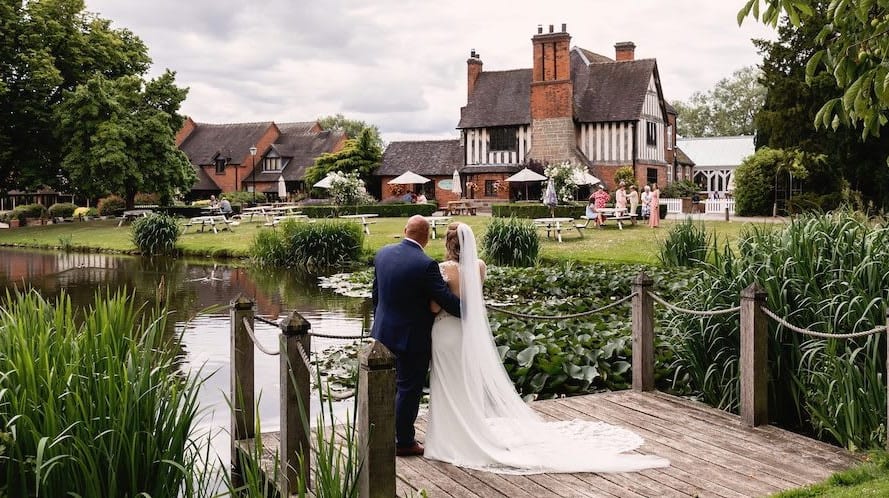 Bride and groom standing on a wooden dock overlooking the pond and historic building at The Moat House, surrounded by lush greenery and wedding guests.