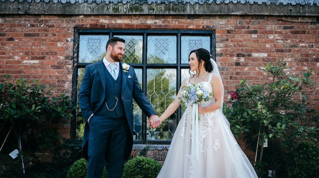 Bride and groom holding hands and smiling outside The Moat House, standing in front of a rustic brick wall with leaded windows and lush greenery.