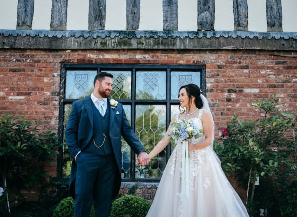 Bride and groom holding hands and smiling outside The Moat House, standing in front of a rustic brick wall with leaded windows and lush greenery.
