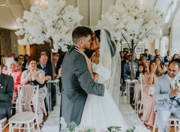 Bride and groom share their first kiss at The Moat House, surrounded by guests and white blossom trees in a beautifully decorated indoor wedding ceremony.