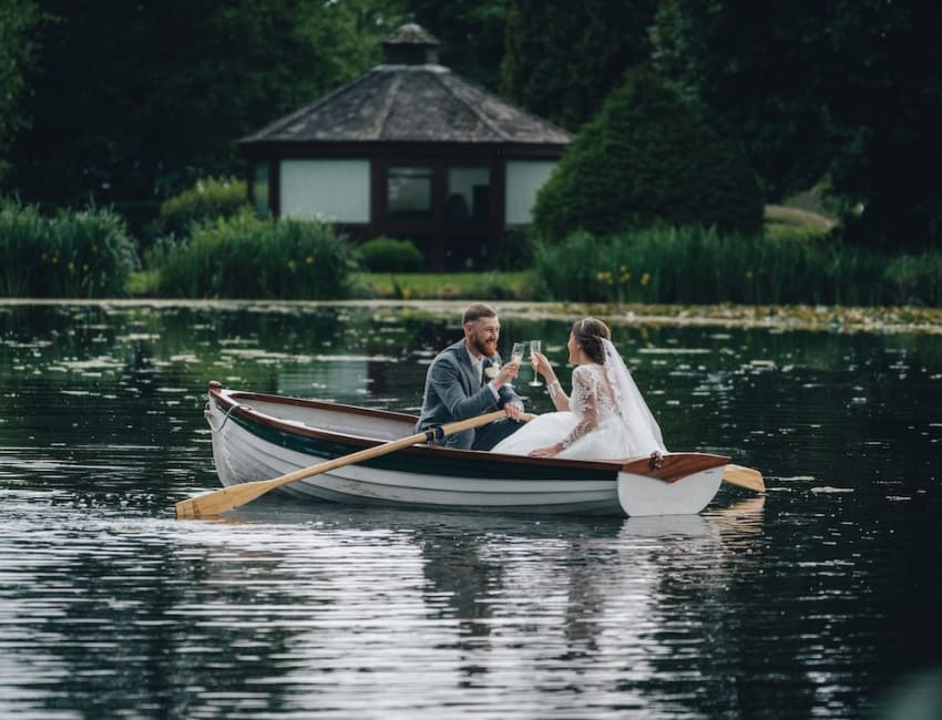 Bride and groom toasting with champagne while sitting in a rowboat on the water at The Moat House, surrounded by lush greenery and a romantic lakeside view.