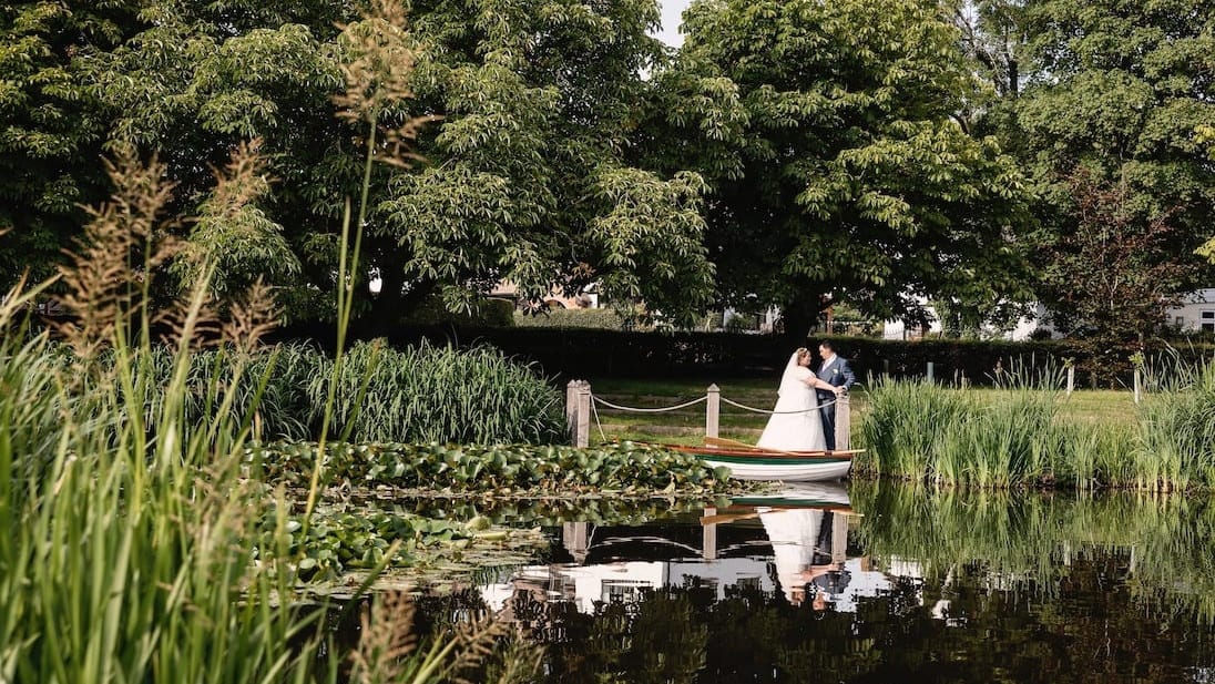 Bride and groom sharing a moment by the water at The Moat House, surrounded by lush greenery with their reflection visible in the calm pond.