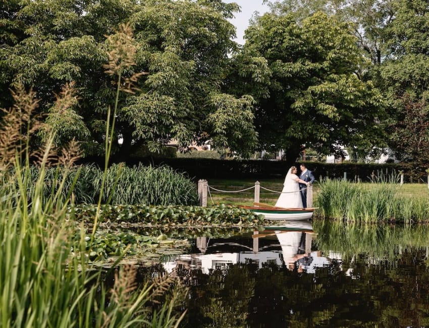 Bride and groom sharing a moment by the water at The Moat House, surrounded by lush greenery with their reflection visible in the calm pond.