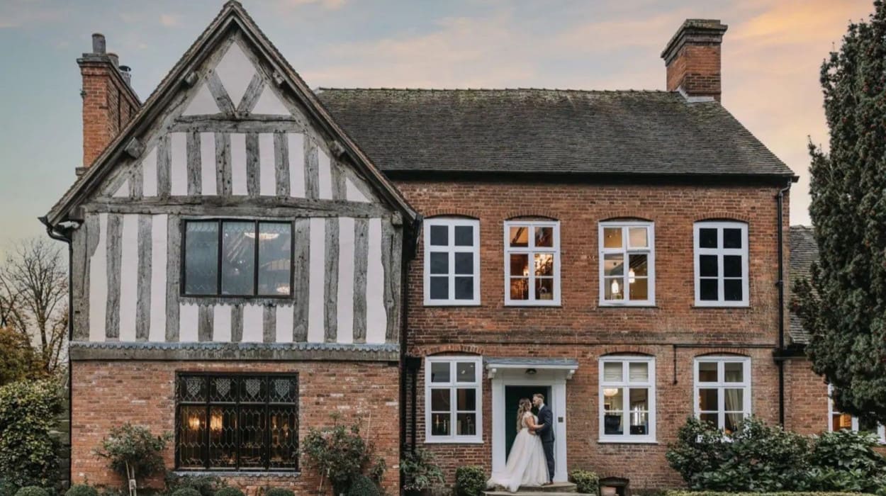 Bride and groom stand in front of The Moat House, a historic red-brick and timber-framed building, captured during golden hour on their wedding day.