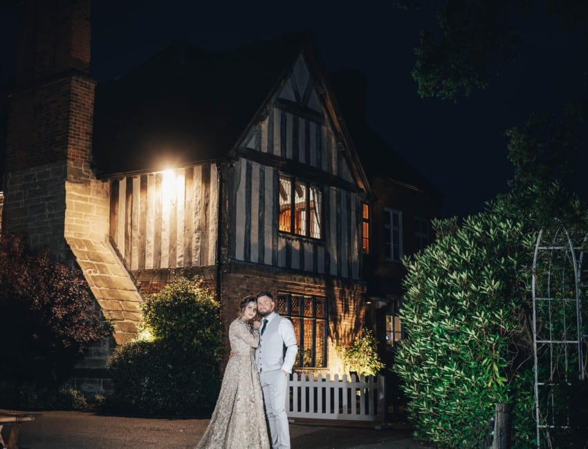 Newlywed couple posing outside The Moat House at night, with warm lighting illuminating the historic timber-framed building and surrounding greenery.