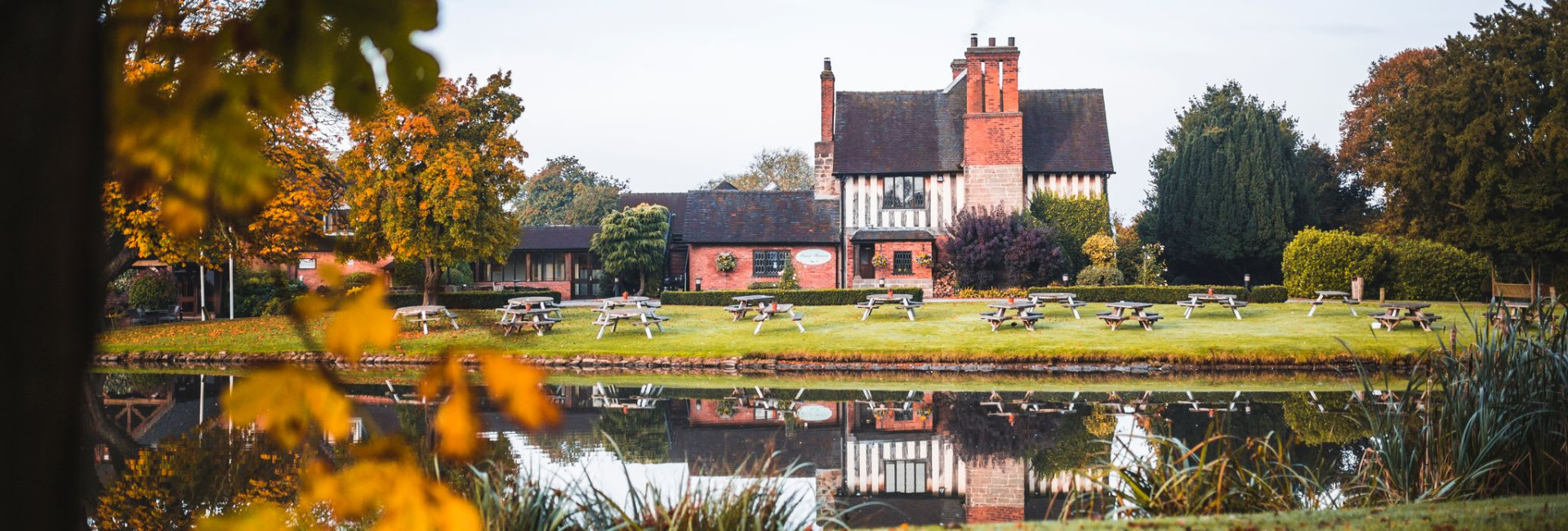 The Moat House in autumn, surrounded by colorful foliage and reflected in a calm pond, with picnic benches on the lawn in front of the historic building.