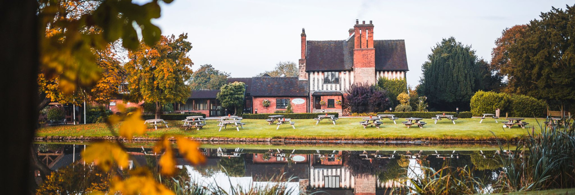The Moat House in autumn, surrounded by colorful foliage and reflected in a calm pond, with picnic benches on the lawn in front of the historic building.