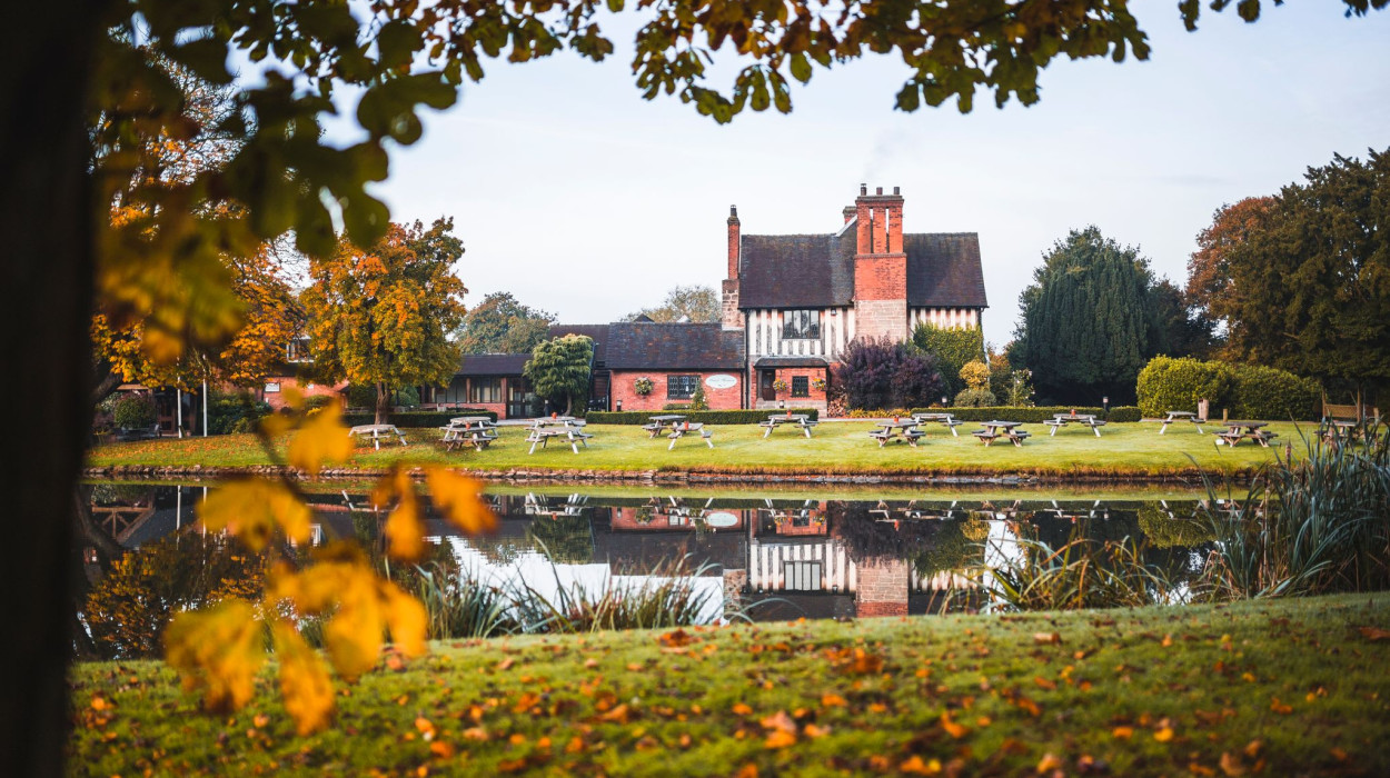 The Moat House in autumn, surrounded by colorful foliage and reflected in a calm pond, with picnic benches on the lawn in front of the historic building.