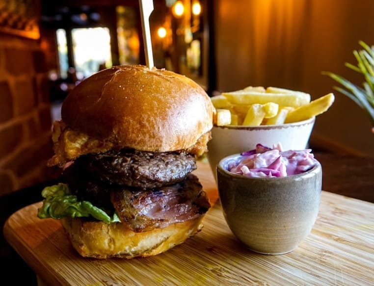 Gourmet burger with lettuce, bacon, and a toasted brioche bun, served with a side of golden fries and purple coleslaw on a wooden board in a cosy restaurant.