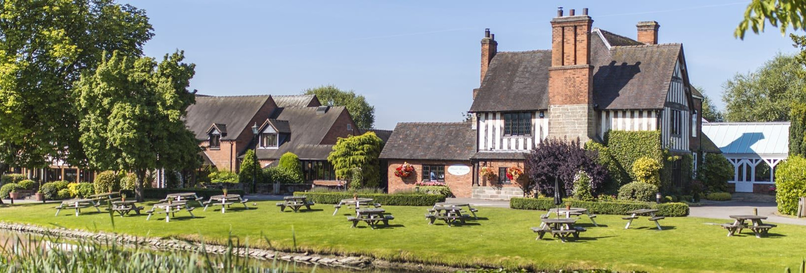 Traditional English country house with timber framing and red brick, surrounded by green lawns, picnic benches, trees, and a pond on a sunny day.