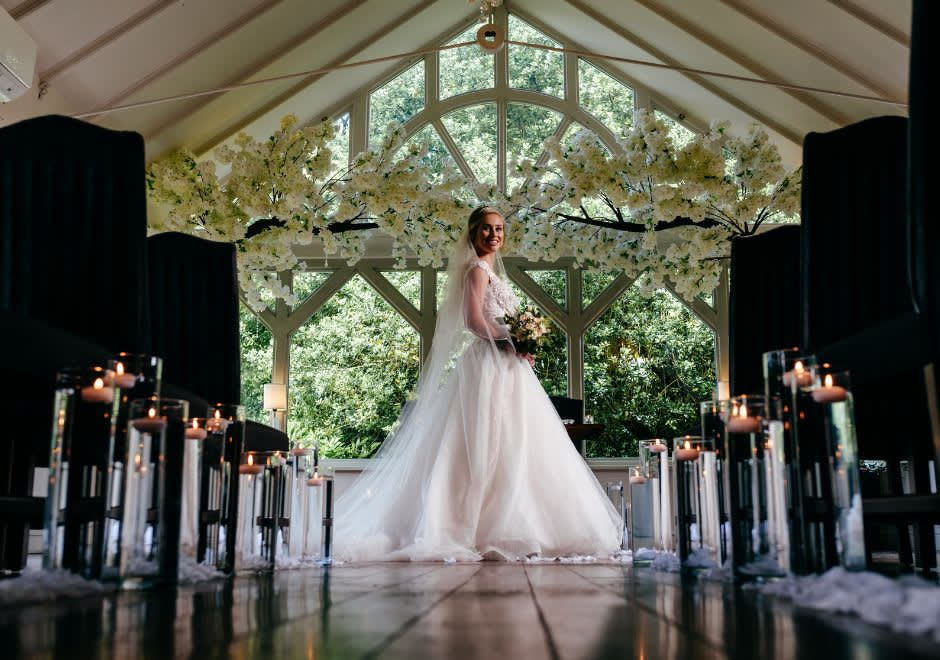 Smiling bride in a flowing white gown holding a bouquet, standing in a candlelit aisle under a floral arch in a bright, windowed ceremony room.
