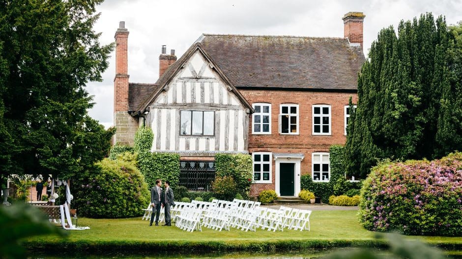 Outdoor wedding ceremony setup with white chairs on a lawn, in front of a Tudor-style brick and timber house, with a pond and lush greenery.