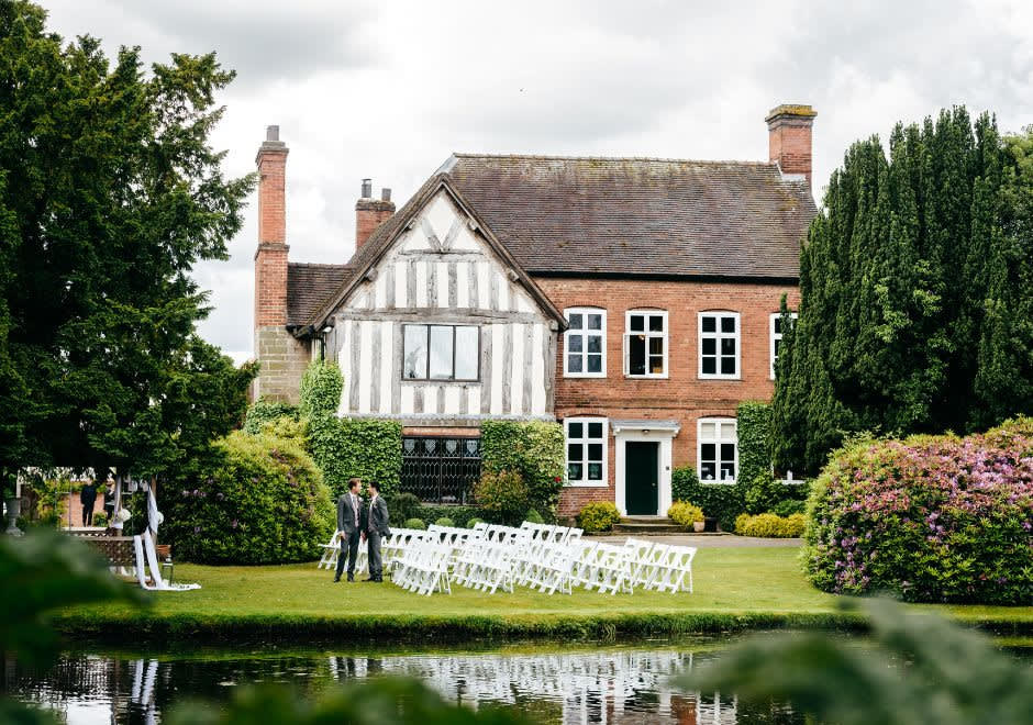 Outdoor wedding ceremony setup with white chairs on a lawn, in front of a Tudor-style brick and timber house, with a pond and lush greenery.