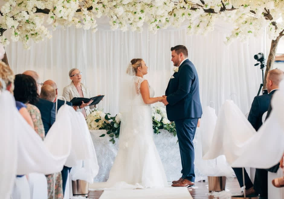 Bride and groom holding hands during their wedding ceremony under a canopy of white flowers, with guests seated and an officiant reading from a book.