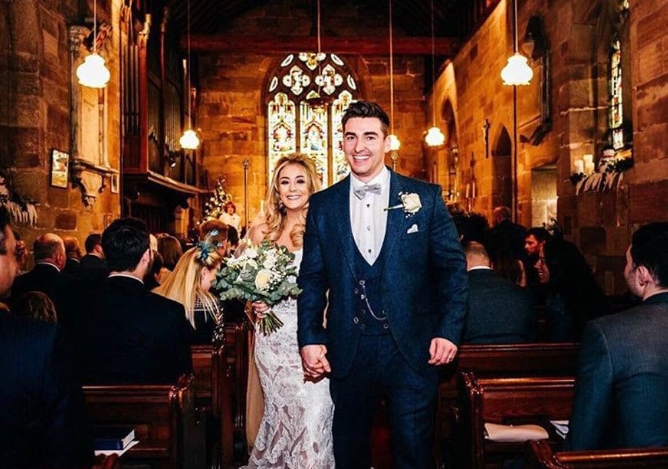 Smiling bride and groom walk hand-in-hand down the aisle of a warmly lit church, surrounded by guests, after their wedding ceremony.