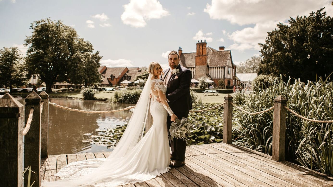 Bride and groom posing on a wooden dock by a lily pond, with a historic half-timbered building and lush greenery in the background on a sunny day.
