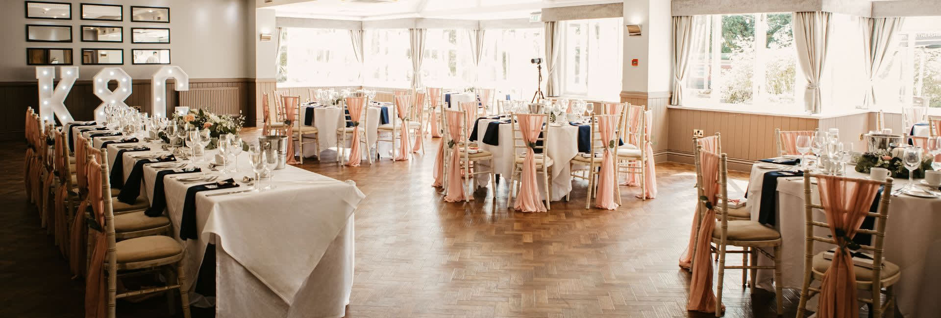 Elegant wedding reception setup with round and long tables, peach chair sashes, navy napkins, floral centerpieces, and large illuminated 