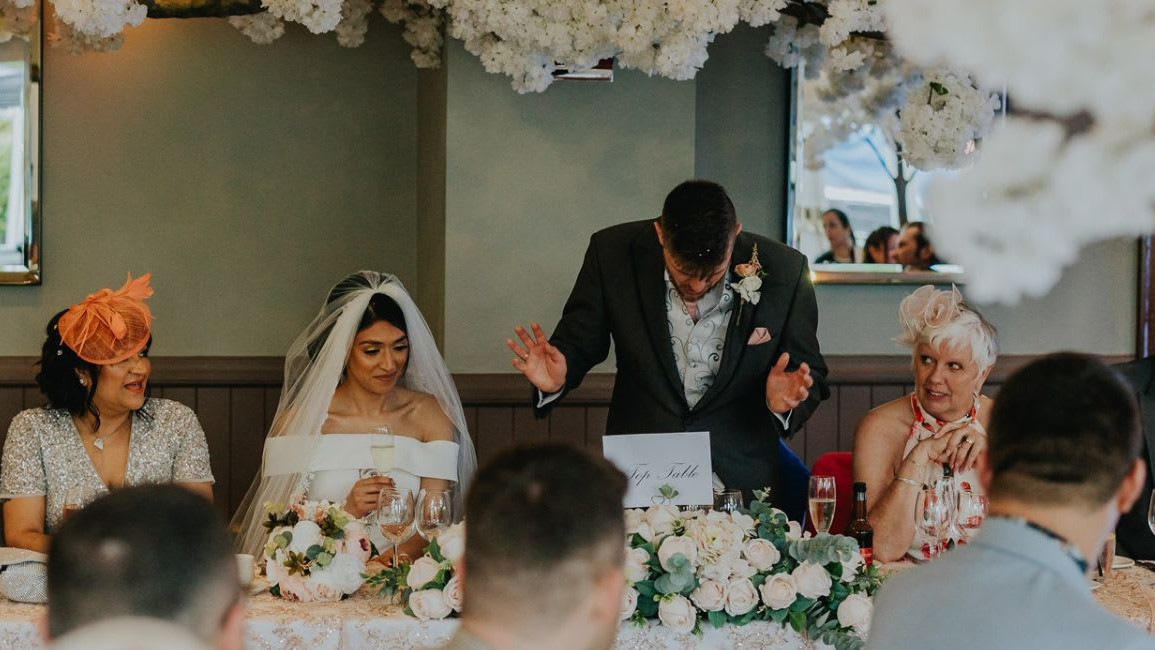 Wedding top table scene with the bride, groom, and guests smiling and reacting as a man gives a speech, surrounded by white floral decorations and mirrors.
