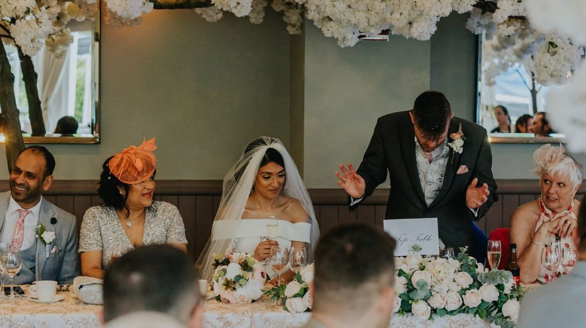 Wedding top table scene with the bride, groom, and guests smiling and reacting as a man gives a speech, surrounded by white floral decorations and mirrors.