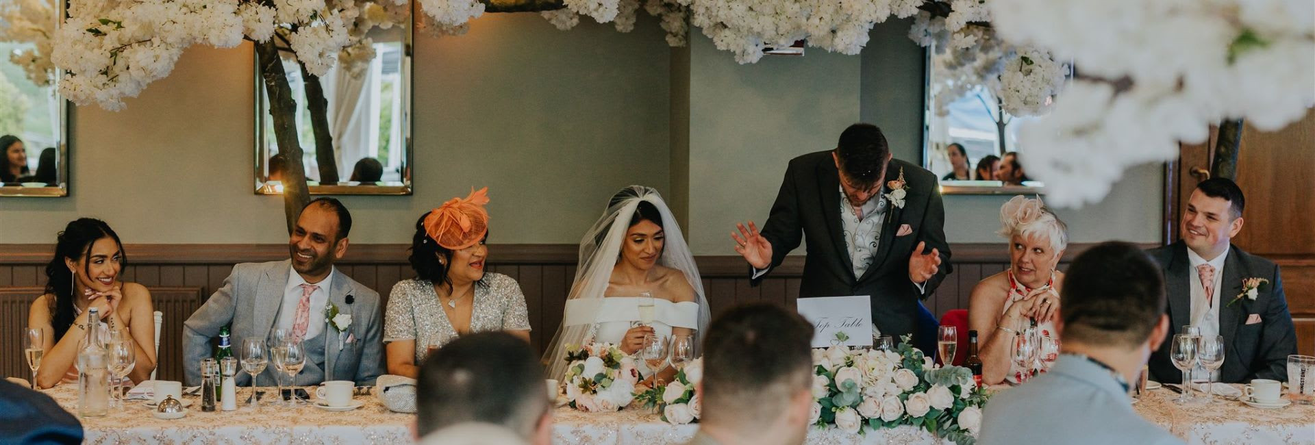 Wedding top table scene with the bride, groom, and guests smiling and reacting as a man gives a speech, surrounded by white floral decorations and mirrors.