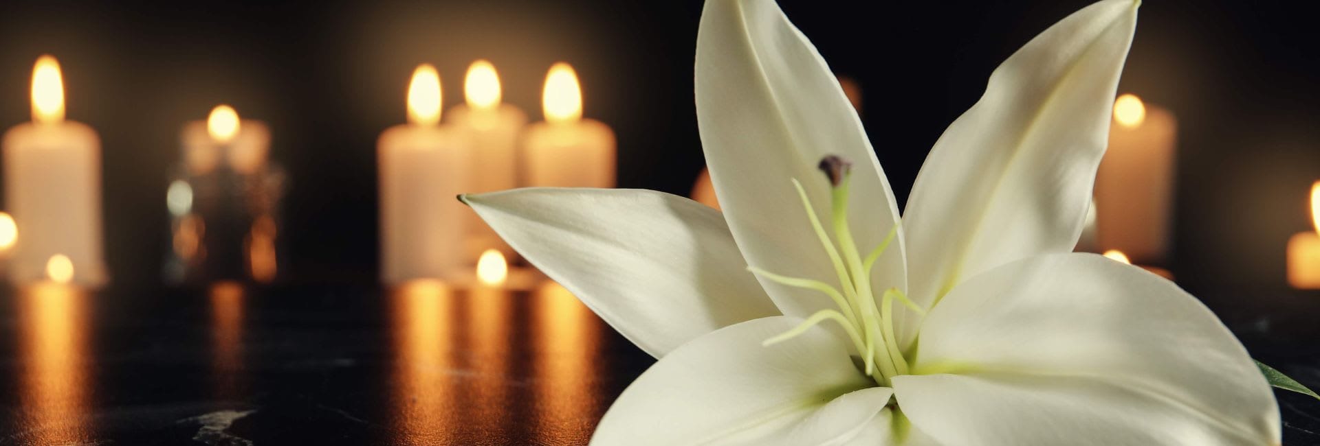 Close-up of a white lily flower with softly glowing candles in the blurred background, creating a calm and serene atmosphere on a dark surface.
