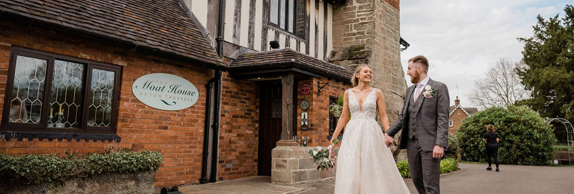A newlywed couple holds hands and smiles outside The Moat House in Acton Trussell, a historic brick and timber building with Tudor-style architecture.