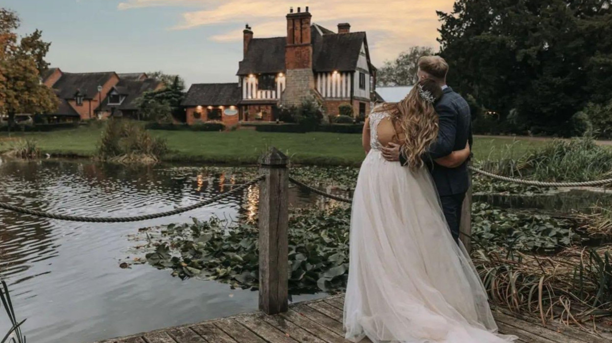 Bride and groom stand on a wooden dock, embracing while gazing at a Tudor-style house across a pond at sunset, surrounded by lush greenery and lily pads.