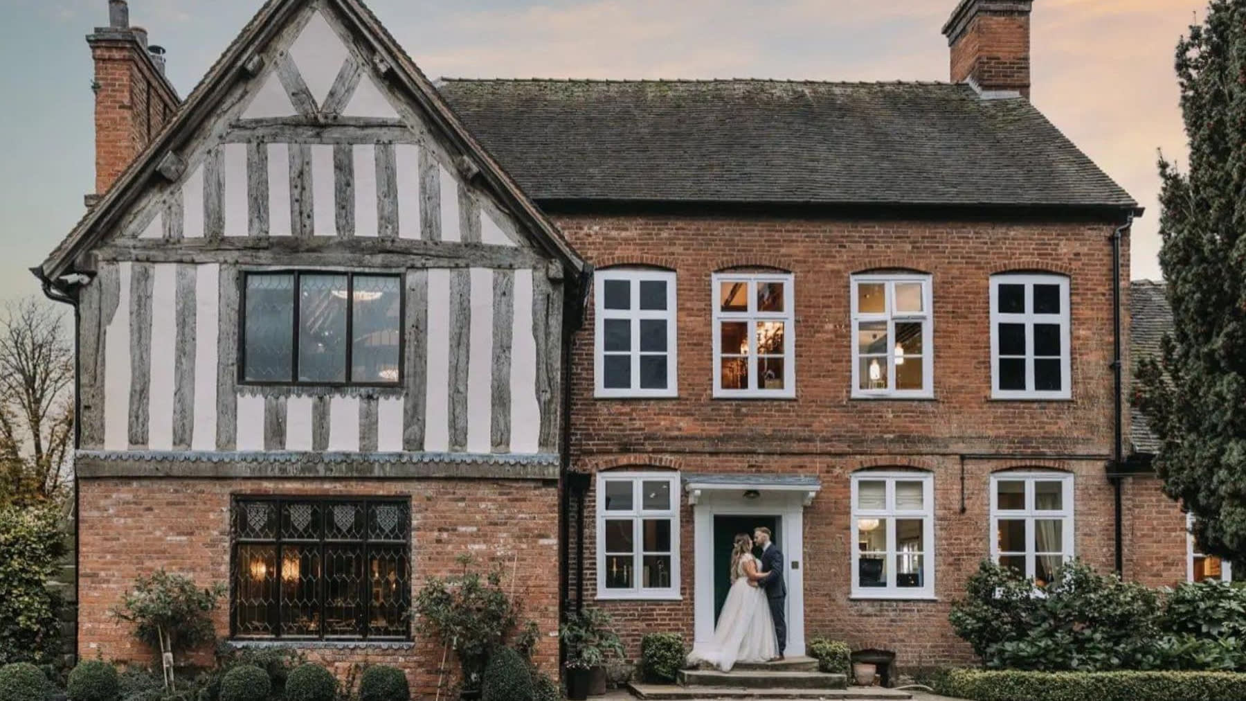 Bride and groom embrace in front of a charming half-timbered and red brick house at sunset, surrounded by neatly trimmed bushes and soft evening light.