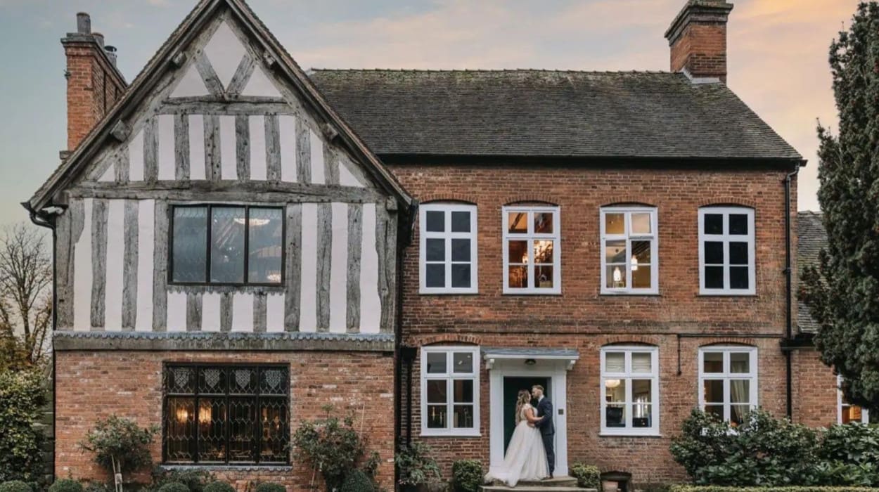 Bride and groom embrace in front of a charming half-timbered and red brick house at sunset, surrounded by neatly trimmed bushes and soft evening light.