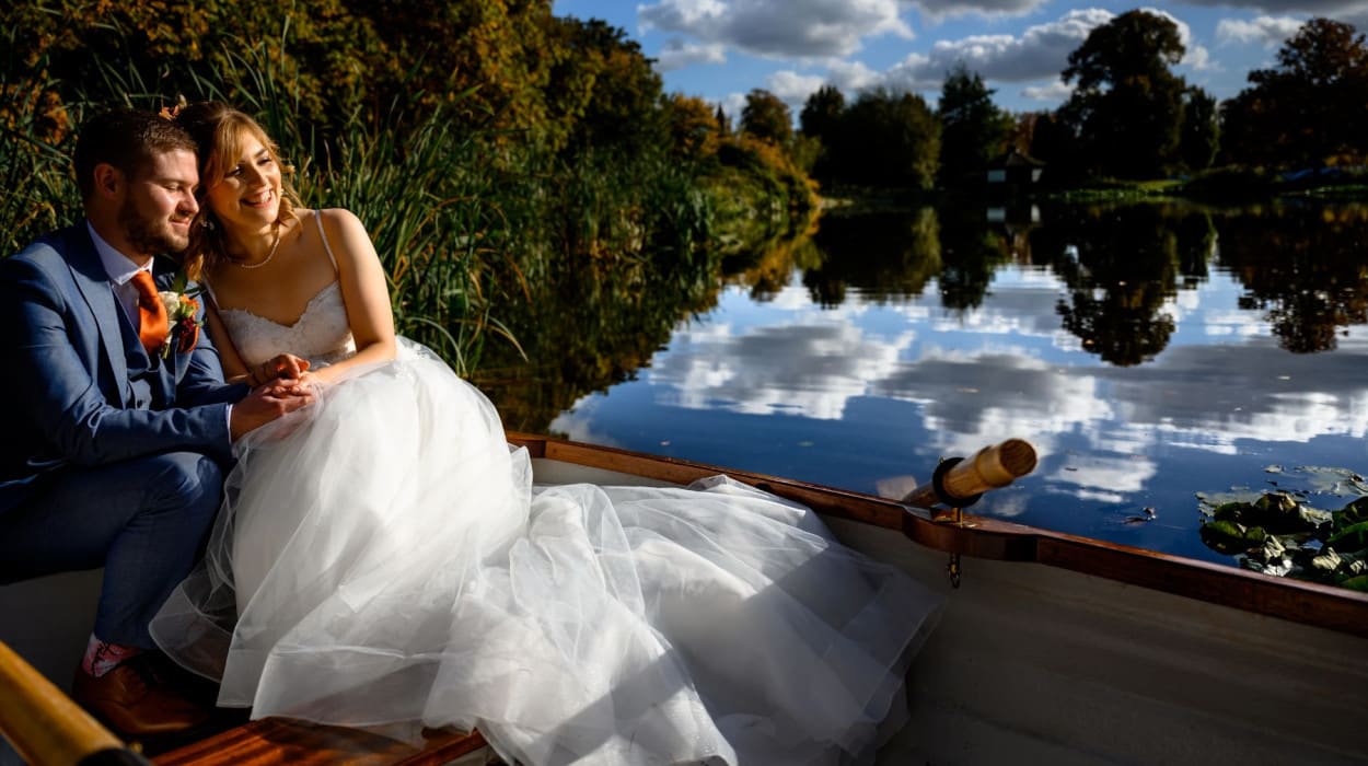 Bride and groom sitting in a rowboat on a calm lake, holding hands and smiling, surrounded by lush greenery and a clear blue sky reflected in the water.
