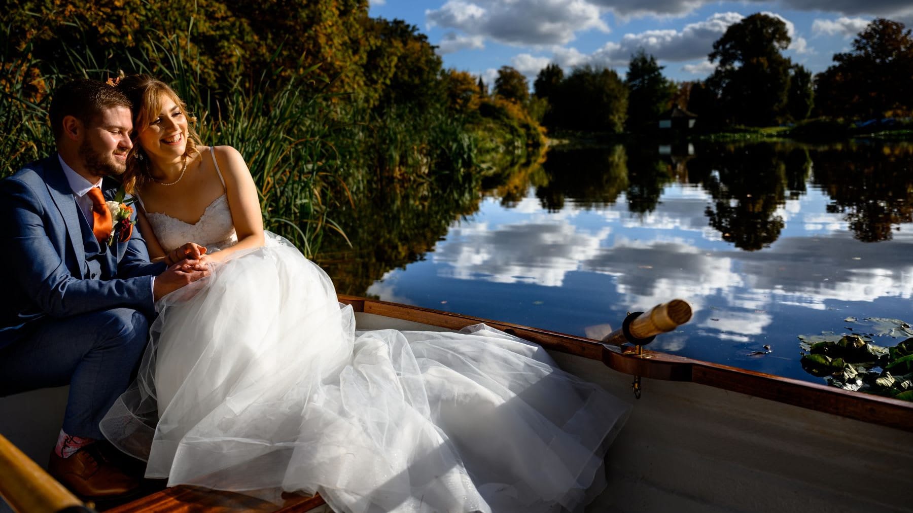 Bride and groom sitting in a rowboat on a calm lake, holding hands and smiling, surrounded by lush greenery and a clear blue sky reflected in the water.