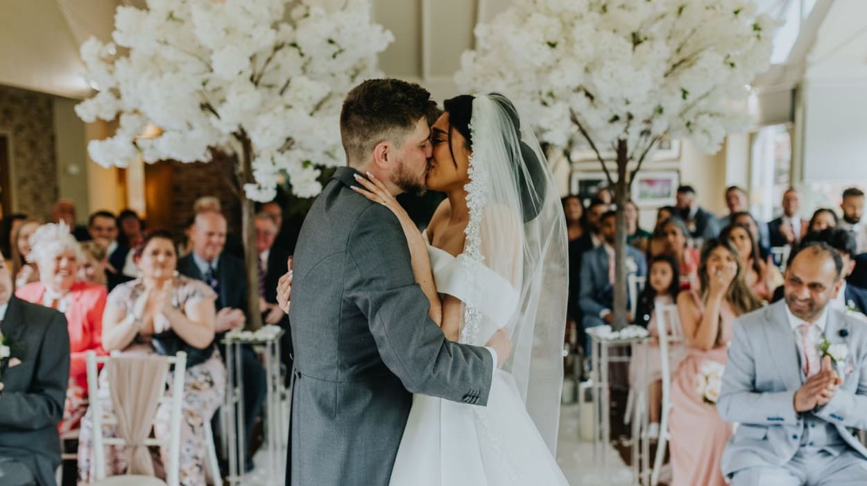 Bride and groom share their first kiss during an indoor wedding ceremony, surrounded by guests and white blossom trees, with joyful expressions all around.