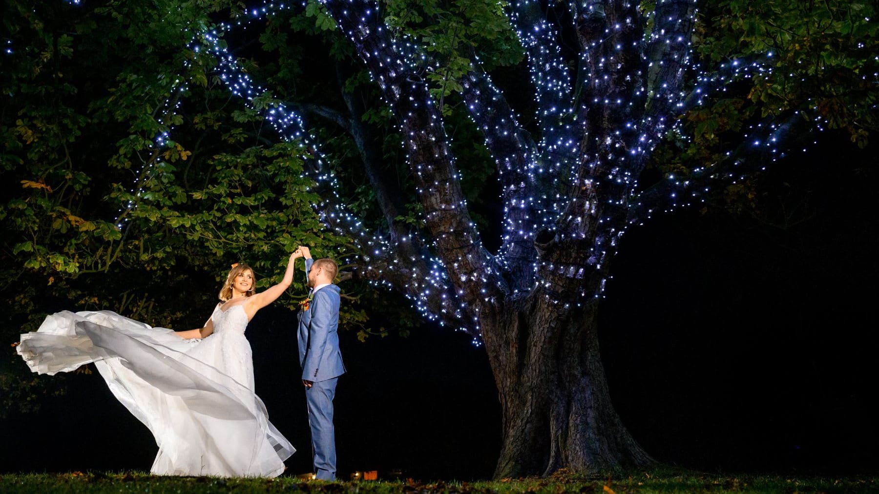 Bride and groom dancing under a large tree wrapped in twinkling fairy lights at night, with the bride's dress elegantly flowing in motion.