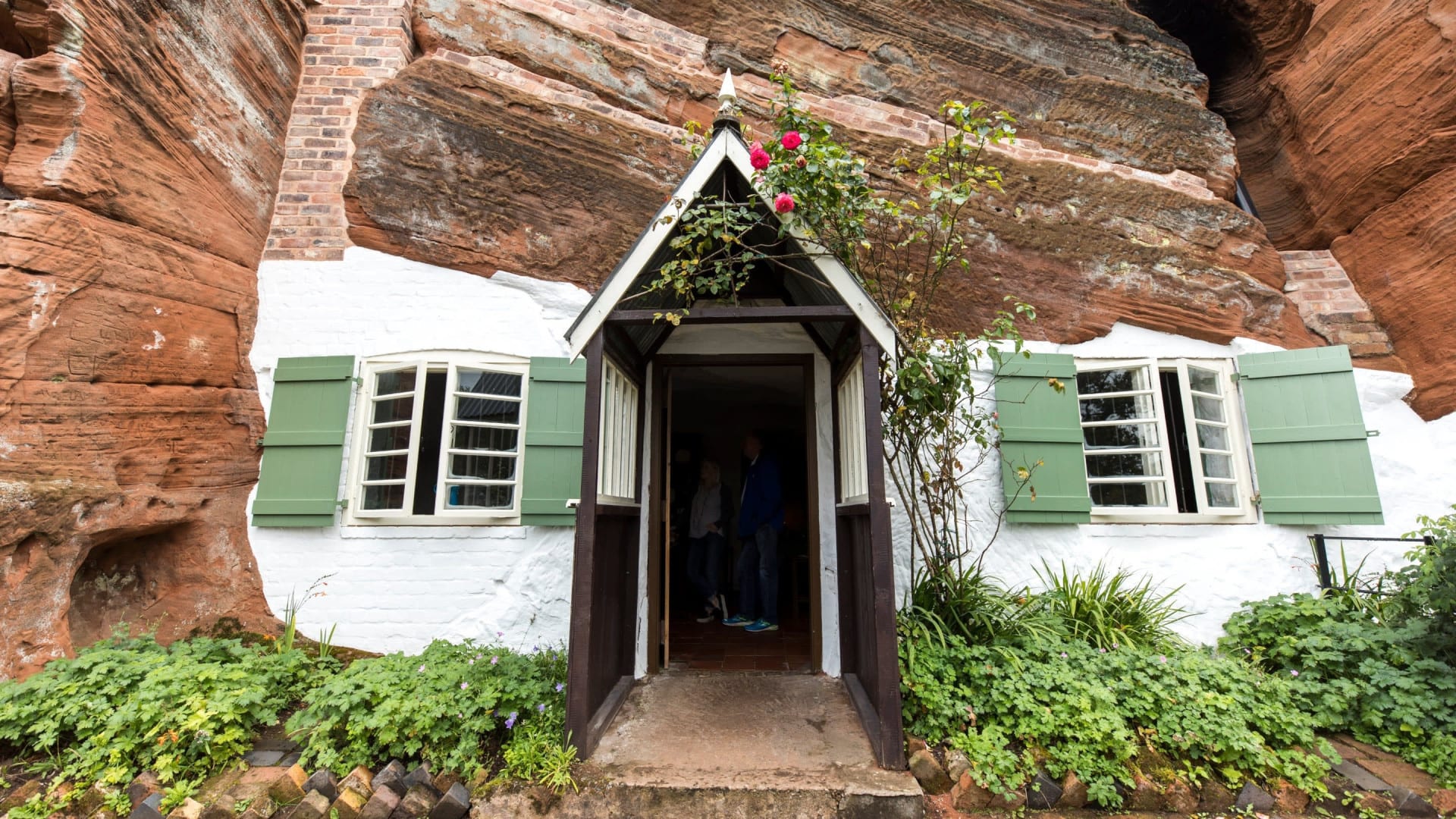 Unique cottage built into red sandstone cliffs, featuring whitewashed brick walls, green shutters, a small porch, and a garden with flowering plants.