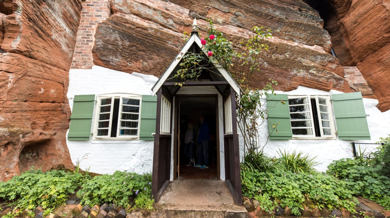 Unique cottage built into red sandstone cliffs, featuring whitewashed brick walls, green shutters, a small porch, and a garden with flowering plants.