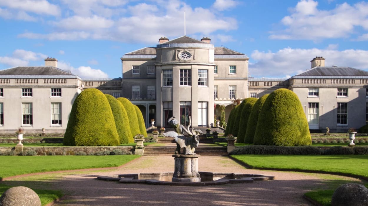Grand historic mansion with symmetrical architecture, topiary trees lining the formal garden, and a central stone fountain under a bright blue sky.